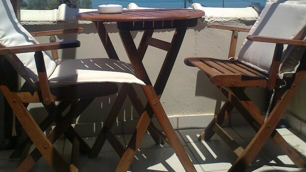 A covered outdoor seating area with two wooden chairs and a small table, overlooking a beach with clear blue water and distant mountains. - Kontogoni Rooms (Elafonisos)