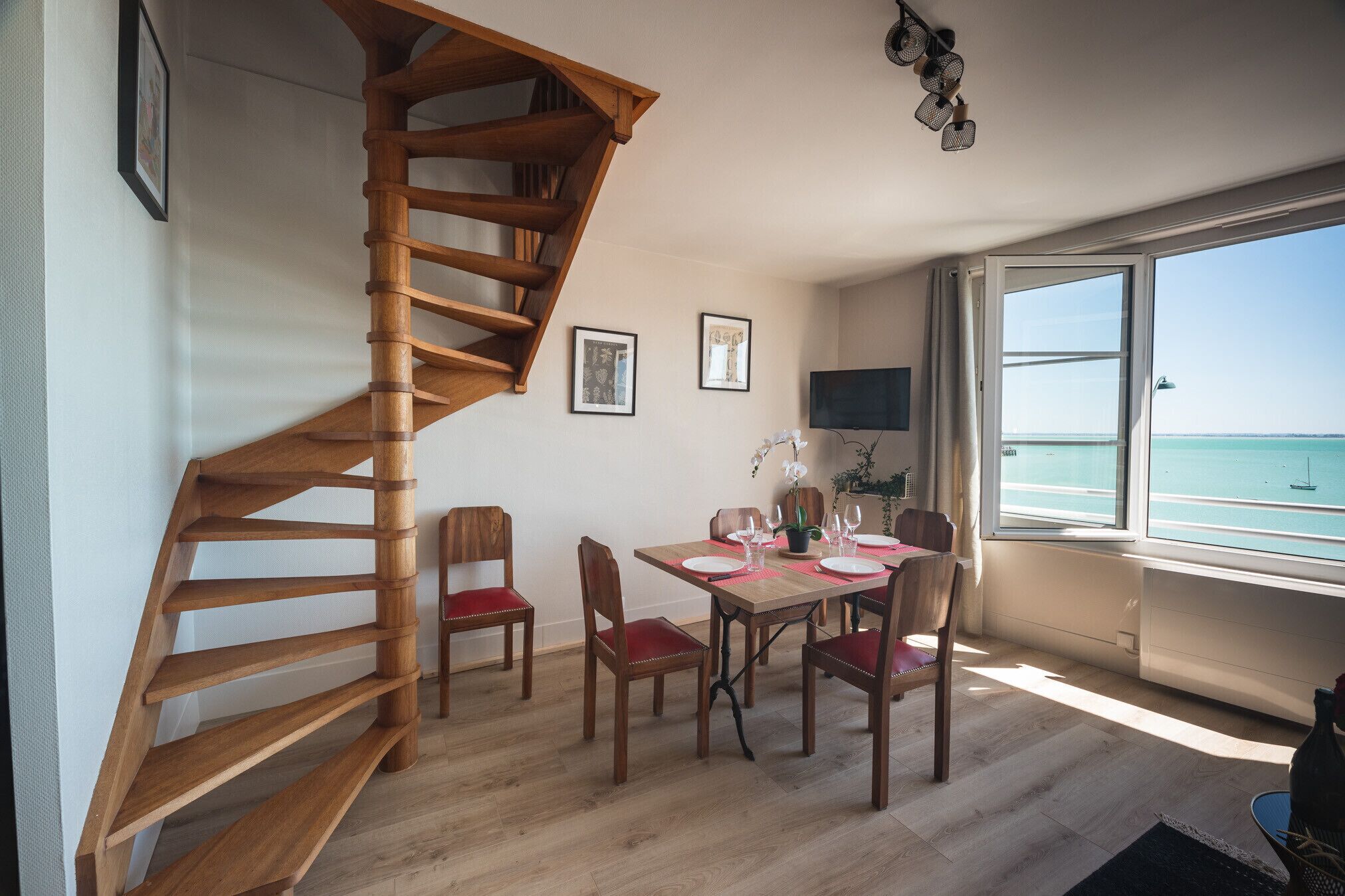 A dining area with a wooden staircase, a table set for four, and a view of the sea through large windows.