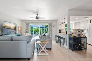A modern living room with a large TV, grey sofas, a coffee table, and a view of a tropical garden through large windows. - Escape to The Sea Turtle's Resort-Like Lanai w/Pool ~Walk to Dining/Shops; Wifi (Marco Island)