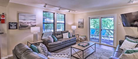 A spacious living room with a leather sofa, a coffee table, and a TV. There are large windows and a view of a deck outside.