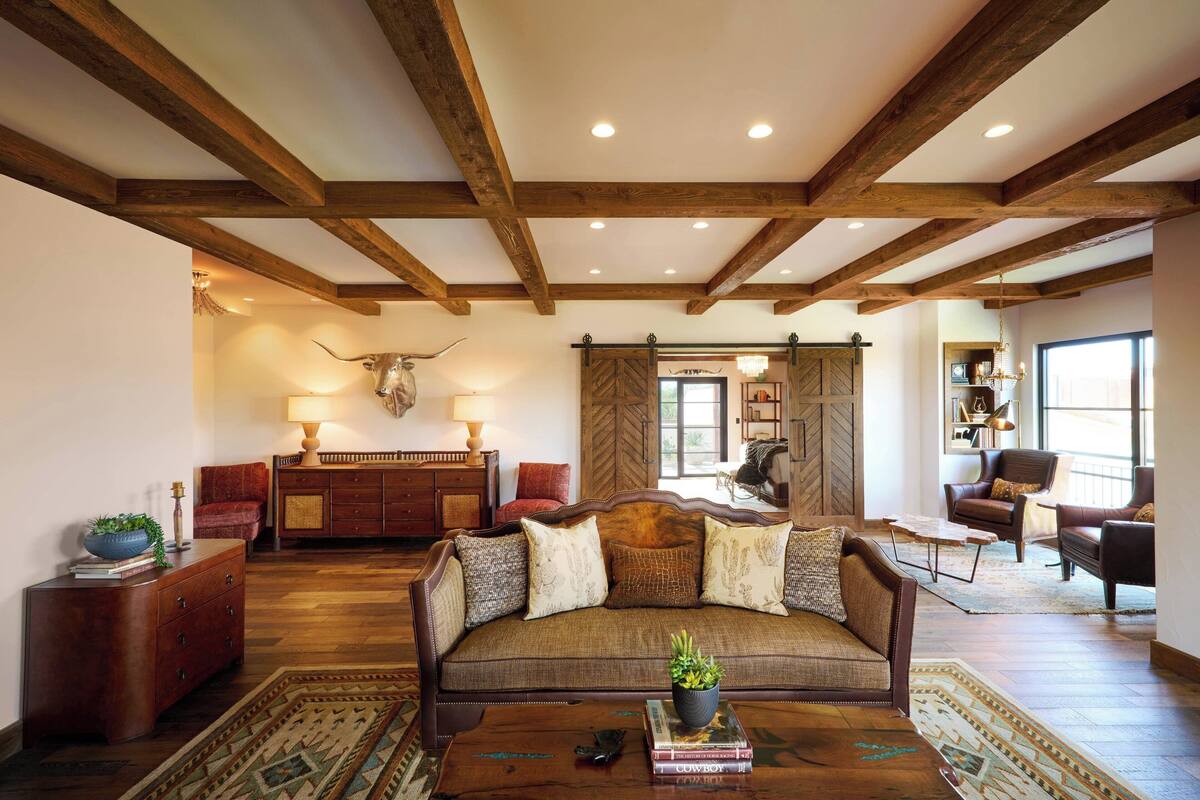 A spacious living room with a wooden ceiling, a sofa with patterned cushions, a coffee table with books, and a decorative antler on the wall.