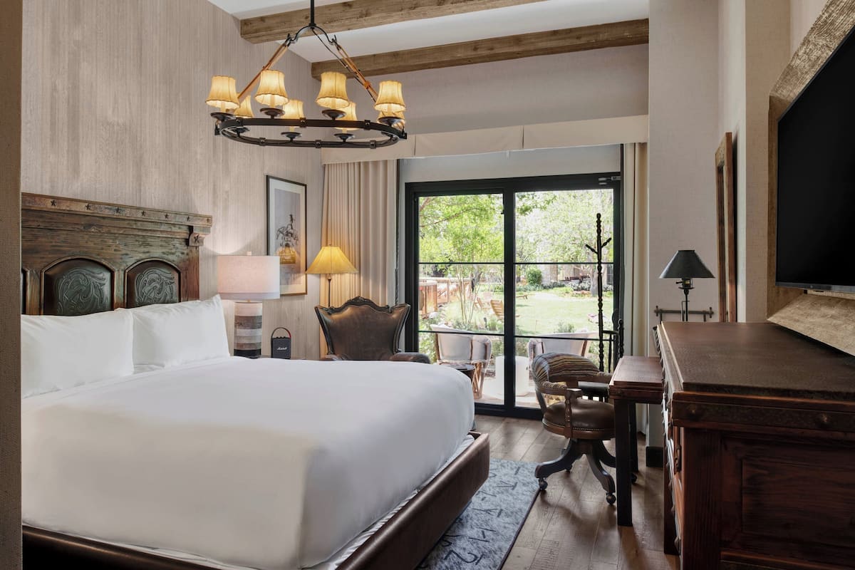 A hotel room with a large bed, a wooden headboard, a chandelier, a desk, and a view of the outdoors.