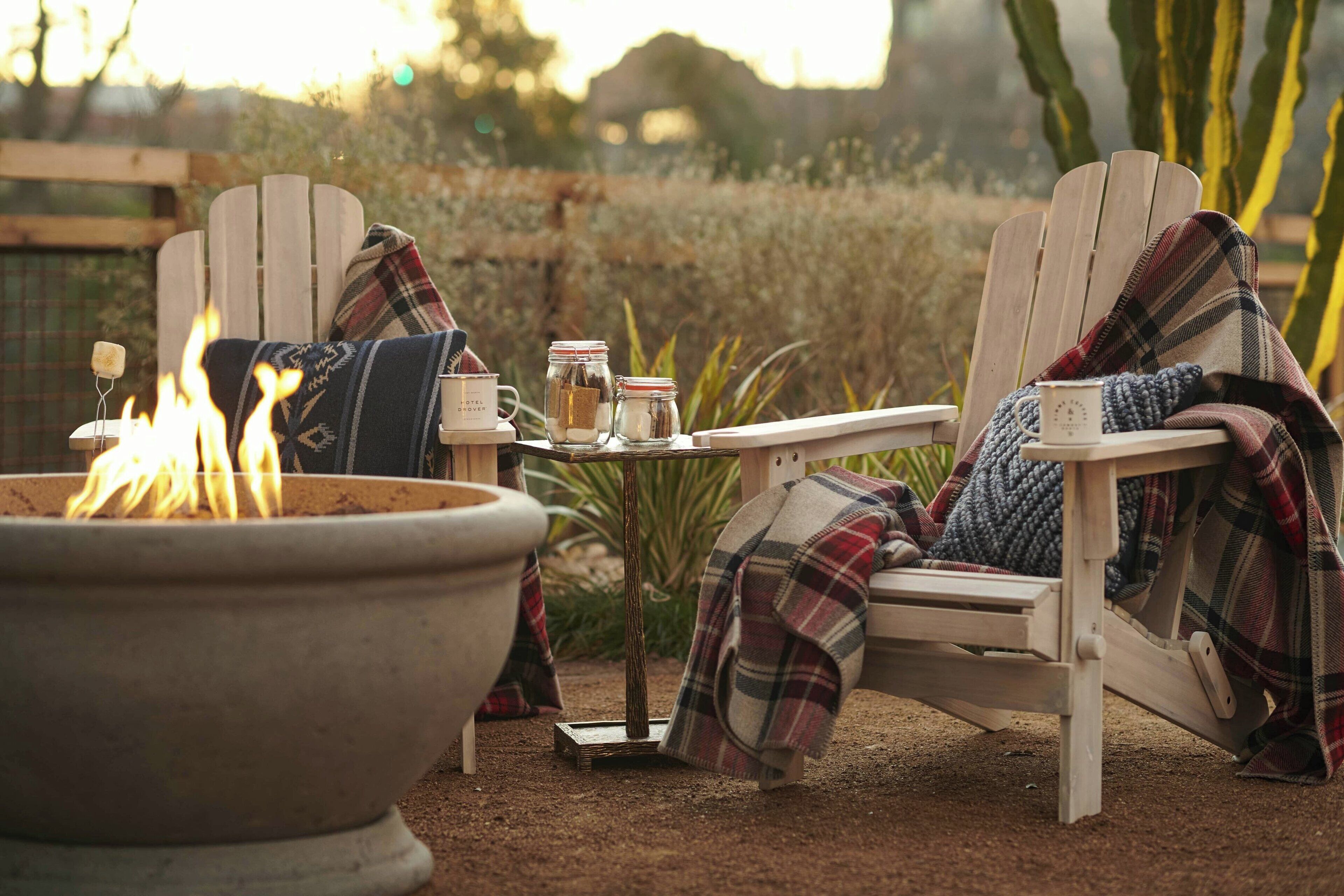 A cozy outdoor seating area with a fire pit, a table with jars and a mug, and a chair with a plaid blanket.