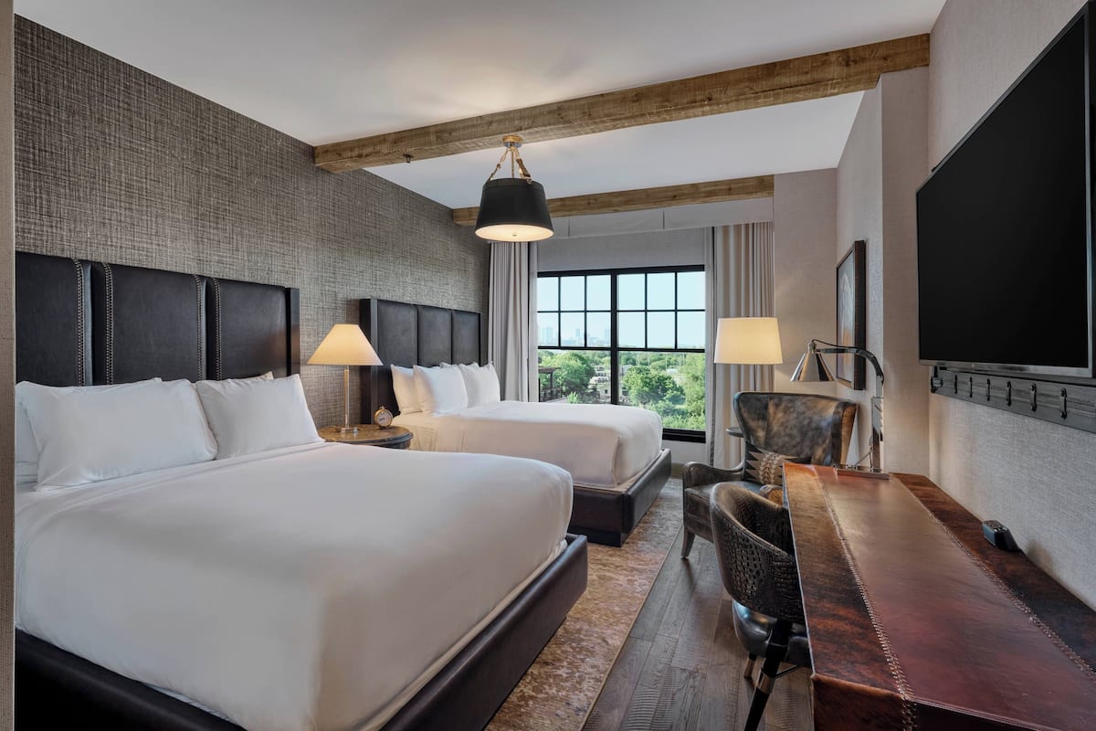 A hotel room with two beds, a wooden ceiling, a large window with a view, a desk with a chair, and a TV mounted on the wall.