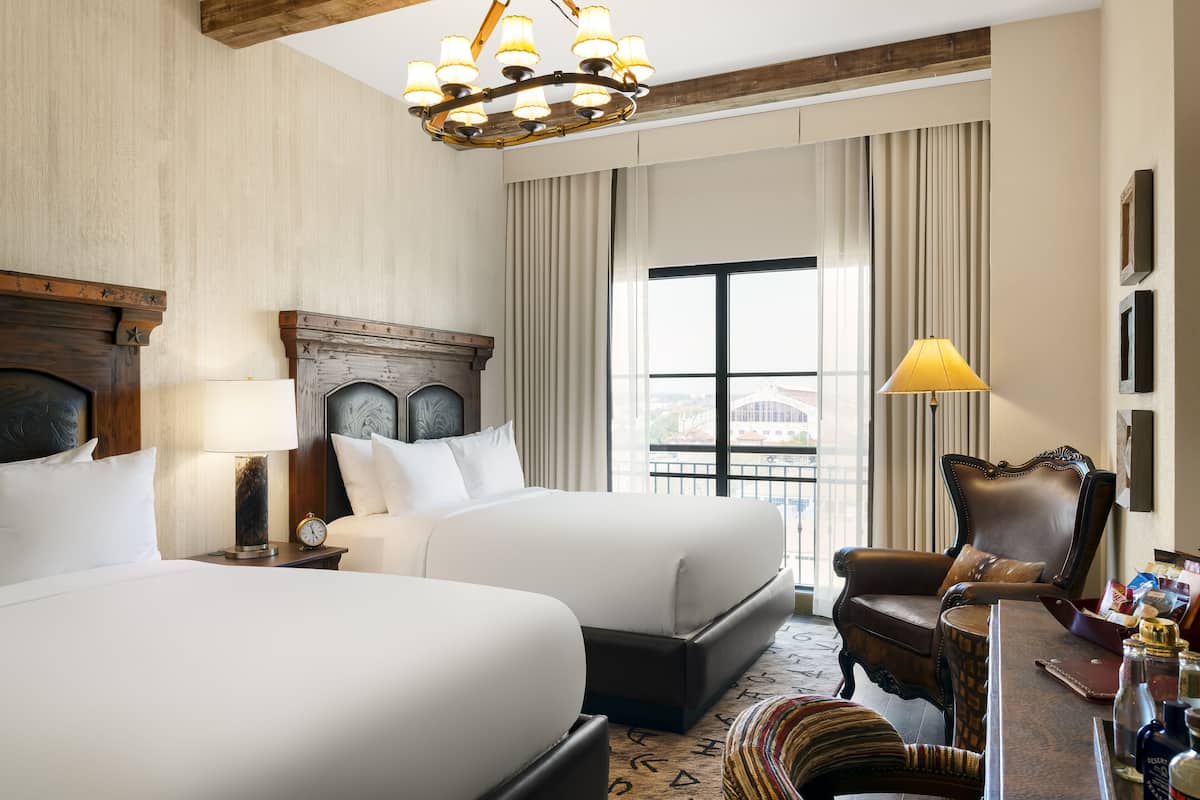 A hotel room with two beds, a wooden headboard, a chandelier, a desk with a lamp, and a view of the city.