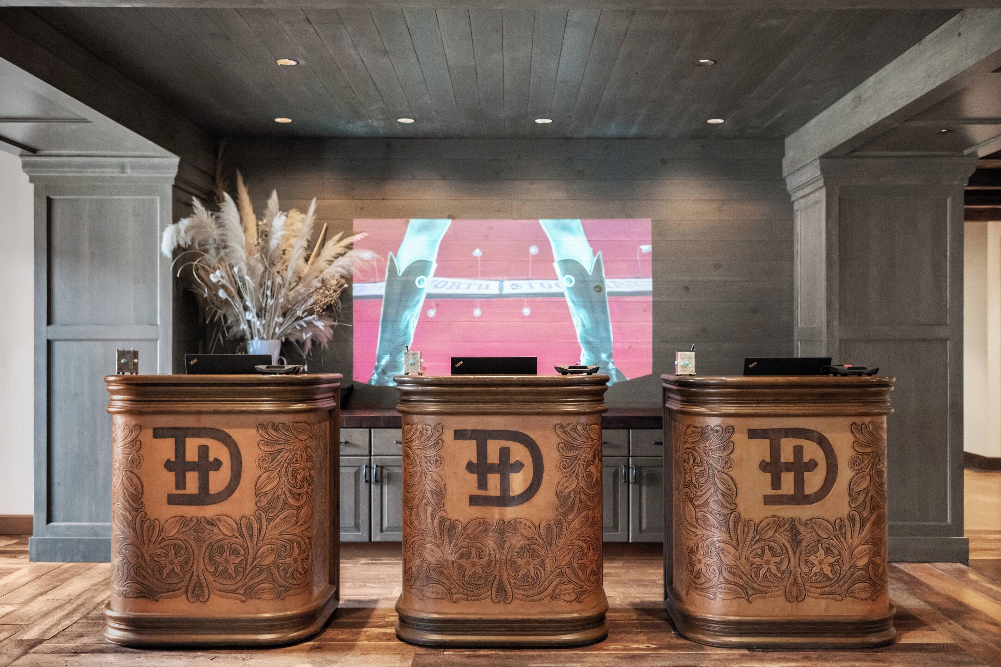 Three ornate, cylindrical containers with a prominent 'D' logo, placed on a wooden floor in a room with a large screen displaying a sports event.