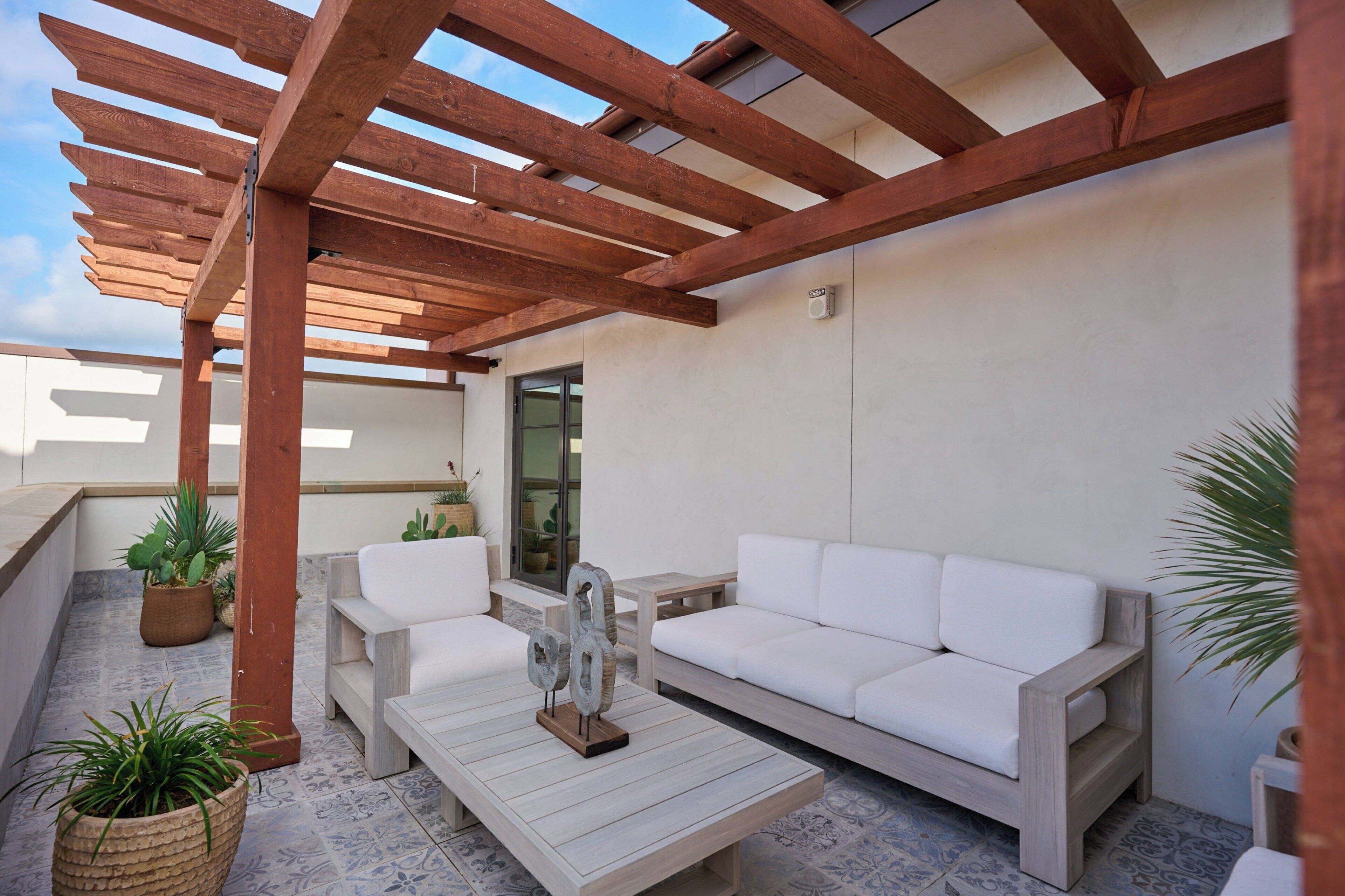 An outdoor patio with wooden pergola, white sofas, a coffee table, and potted plants.