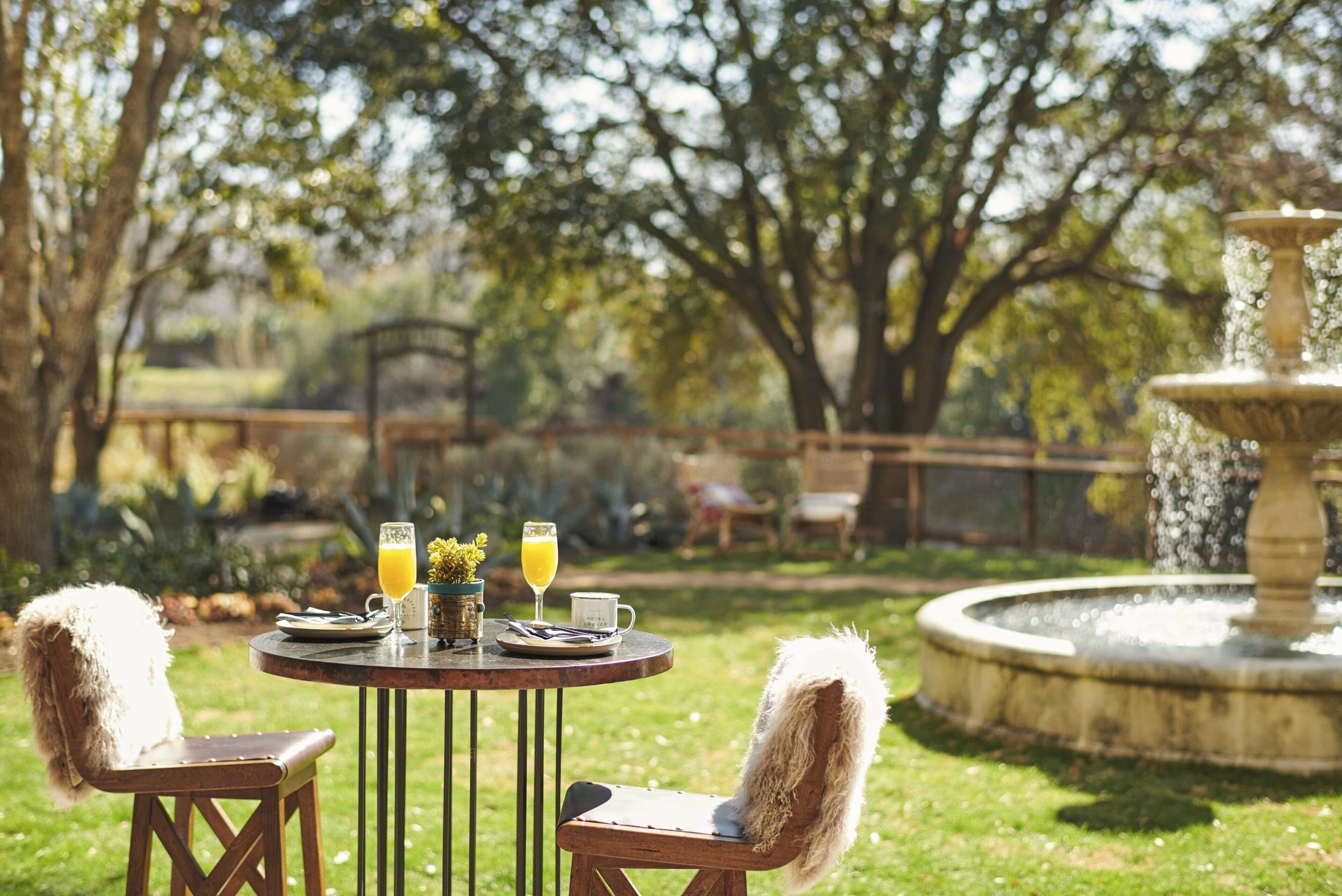 An outdoor dining area with a round table, two chairs, a fountain, and a garden in the background.
