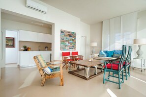 Una sala de estar moderna con una mesa de centro de madera, sillas a juego y un sofá con cojines azules. Un cuadro abstracto y colorido adorna la pared, y se ve una cocina al fondo.