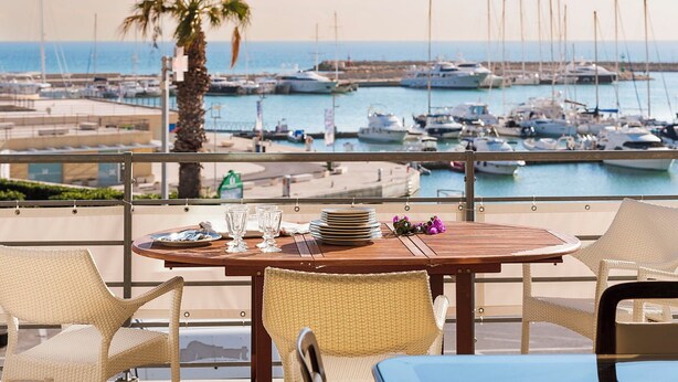 Una mesa de comedor con platos, vasos y cubiertos en un balcón con vistas a un puerto deportivo con barcos.