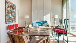 Una sala de estar con una mesa de centro de madera, sillas rojas y azules, un sofá con cojines estampados y un ventanal con vistas.