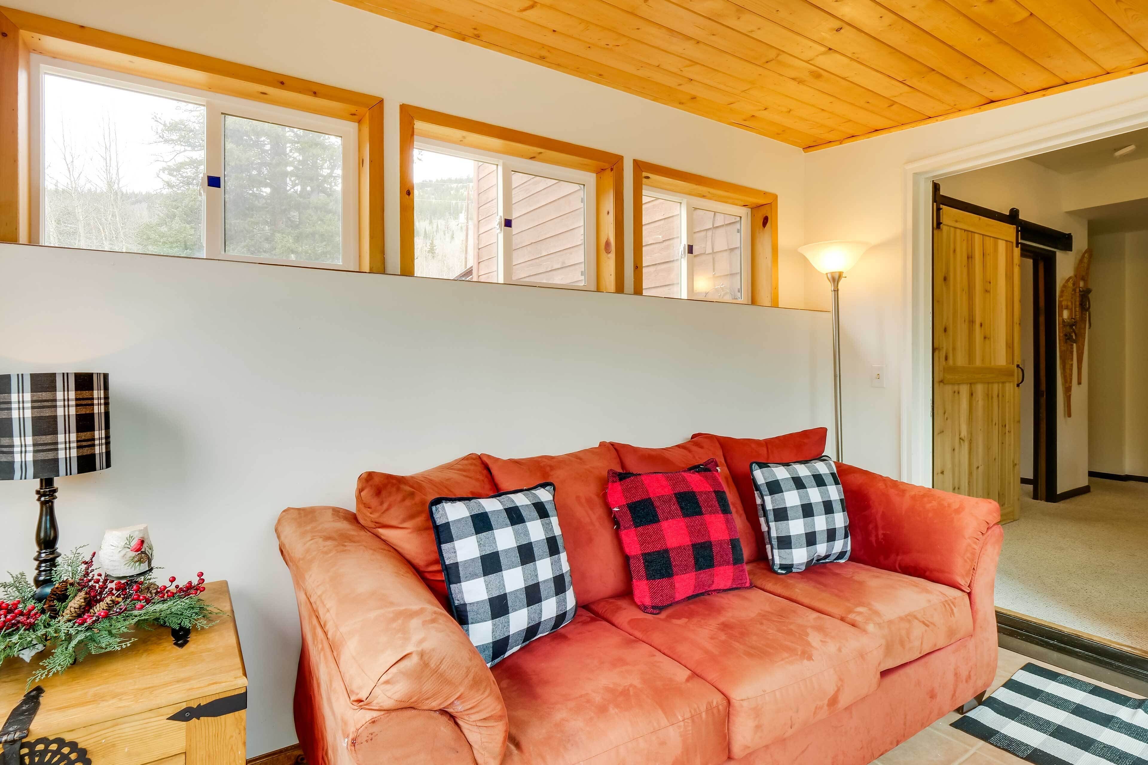 A cozy living room with a plush orange sofa, checkered pillows, a wooden side table with a lamp, and a window with a view of trees.