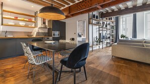 A modern kitchen with wooden beams, a dining table with chairs, and a sofa in a living area with large windows.