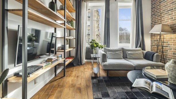 A modern living room with a wooden floor, a grey sofa, a TV, and a bookshelf.
