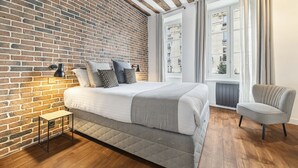 A bedroom with a brick wall, a bed with a grey quilt, a small table, a chair, and a window with curtains.
