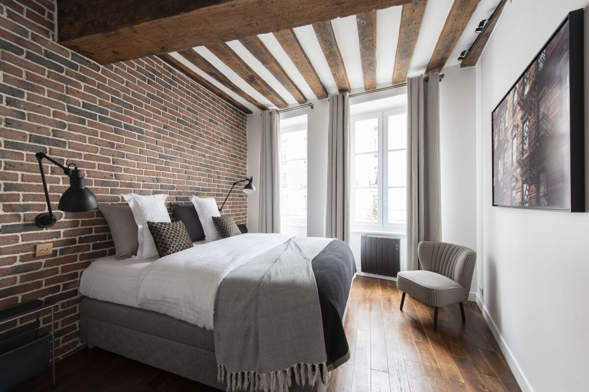 Ein Schlafzimmer mit Backsteinwand, einem Bett mit grau-weißer Bettdecke, einem Sessel und einem großen Fenster mit Vorhängen.