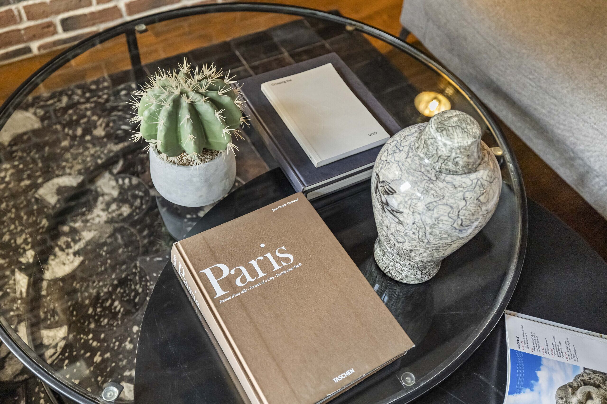 A glass table with a book titled 'Paris', a potted cactus, a decorative vase, and a candle.
