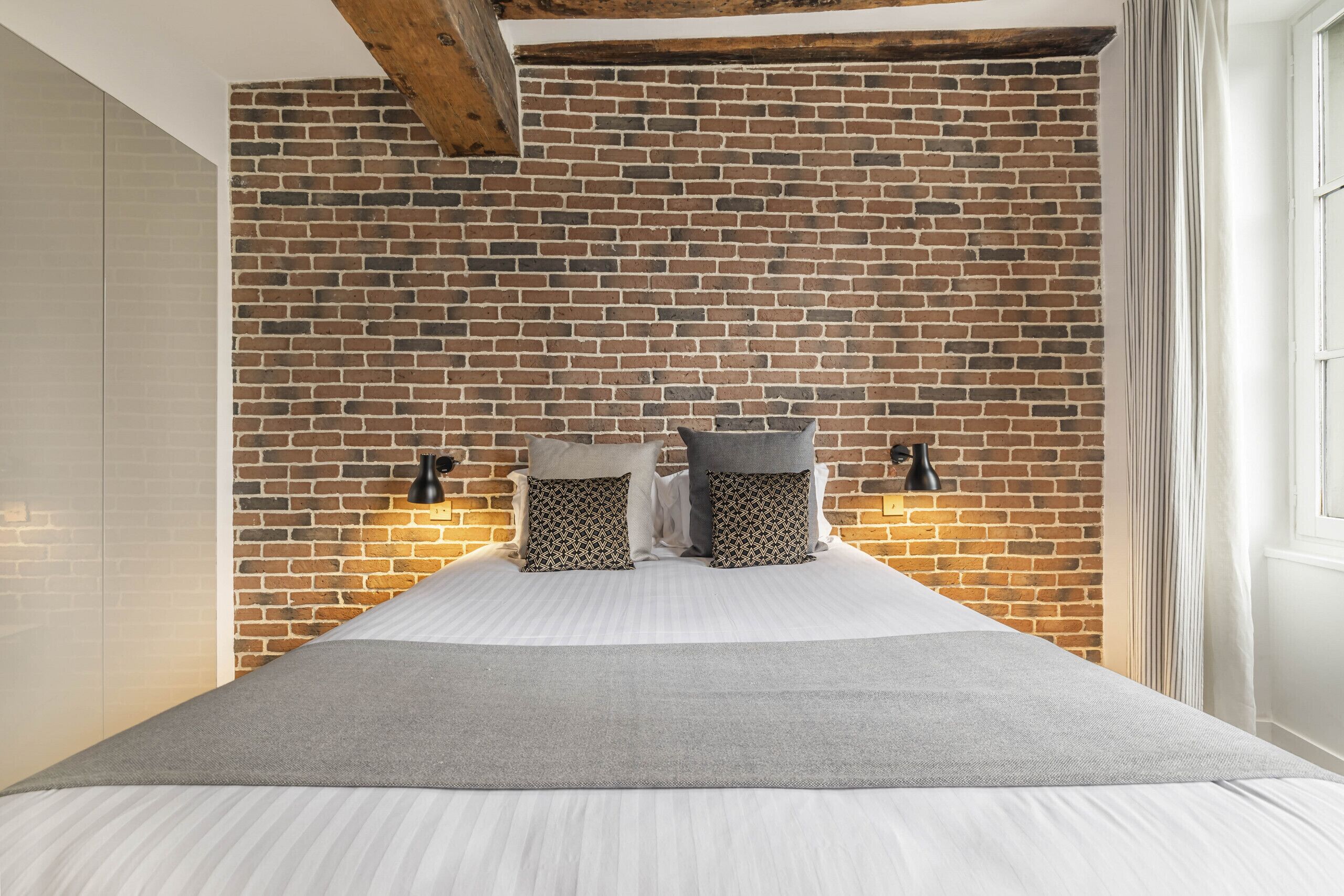 A bed with a grey blanket, two patterned pillows, and a brick wall behind.