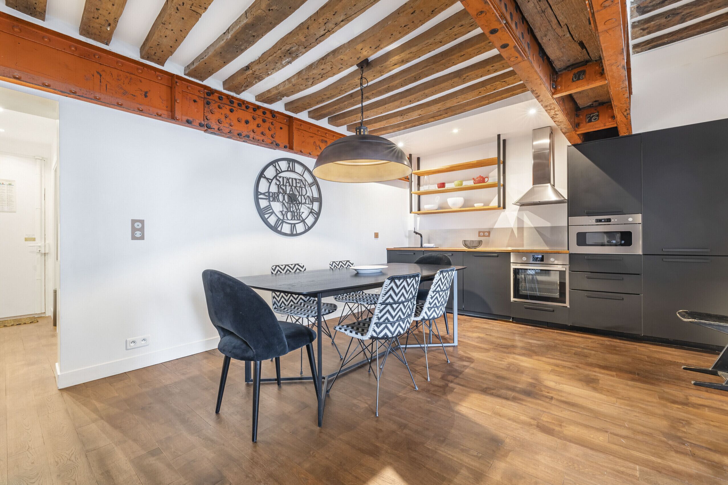 A modern kitchen with wooden beams, a dining area with a black and white patterned table, and a clock on the wall.