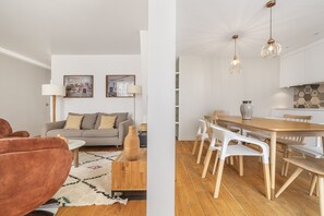 A modern living room with a sofa, coffee table, and artwork on the wall. Adjacent is a dining area with a wooden table and chairs.
