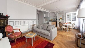 Un salon moderne avec un canapé gris, une chaise en bois, une petite table ronde et un tapis rouge. On aperçoit une cuisine à l’arrière-plan, avec des placards et un coin repas.