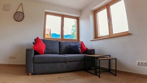 Een kamer met een grijze bank, twee ramen, een kleine tafel en een wandklok.