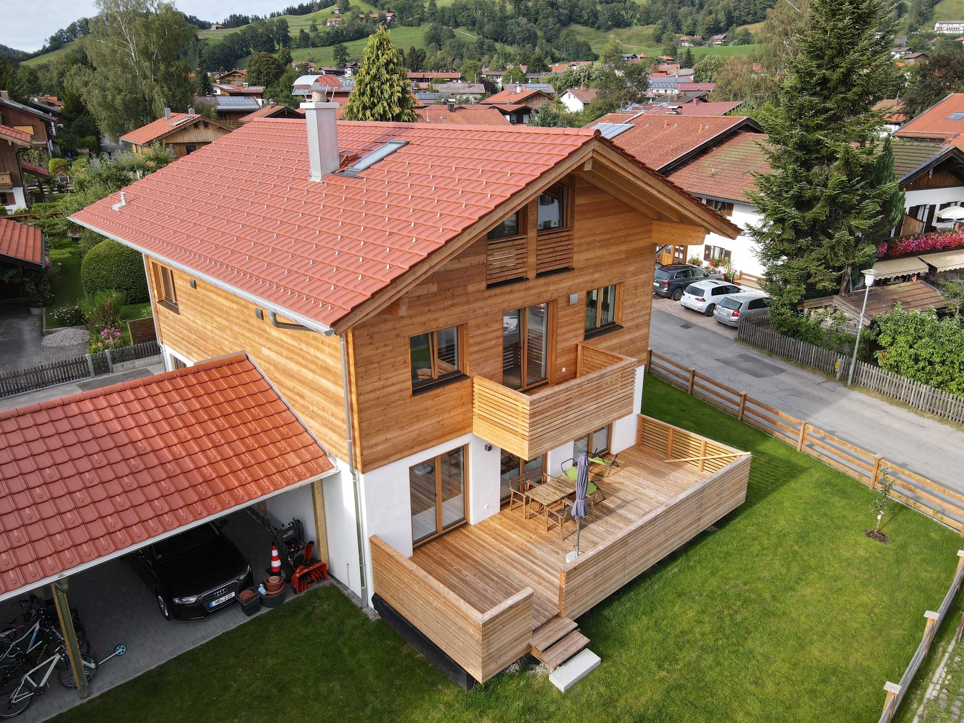 Ein modernes, zweistöckiges Haus mit rotem Ziegeldach, einer Holzterrasse und einer Garage.