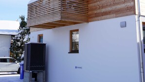 Een houten huis met twee verdiepingen, een balkon, een zichtbaar raam en een klein buitengebied met een vuilnisbak.