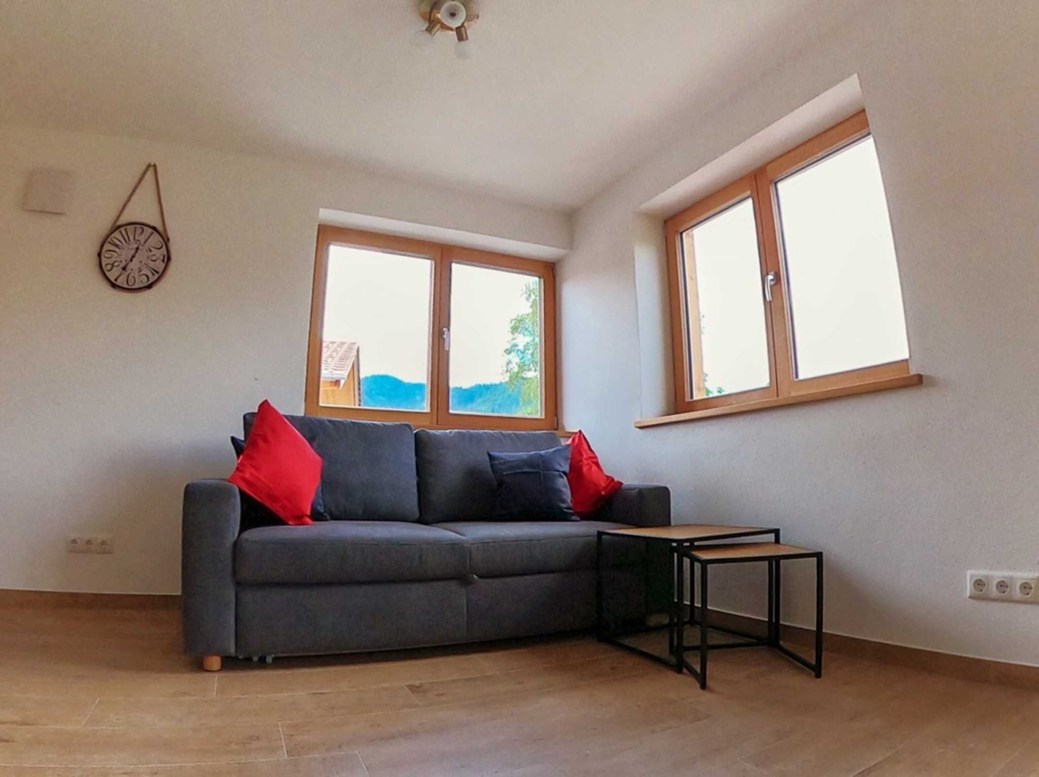 Ein Zimmer mit grauem Sofa, zwei Fenstern, einem kleinen Tisch und einer Wanduhr.