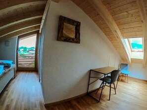 A loft-style room with a sloped ceiling, a small table with chairs, a bed, and a large window.
