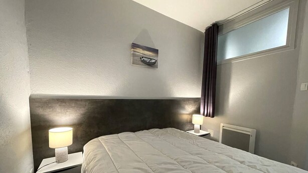 A hotel room with a bed, two bedside lamps, a nightstand, a window with curtains, and a wall-mounted picture. - Well-Equipped Apartment, 500m from Beach and Shops in Perros-Guirec (Perros-Guirec)