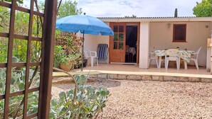 Un petit bâtiment d’un seul étage doté d’un patio comprenant une table et des chaises, un parasol bleu et des plantes en pot.