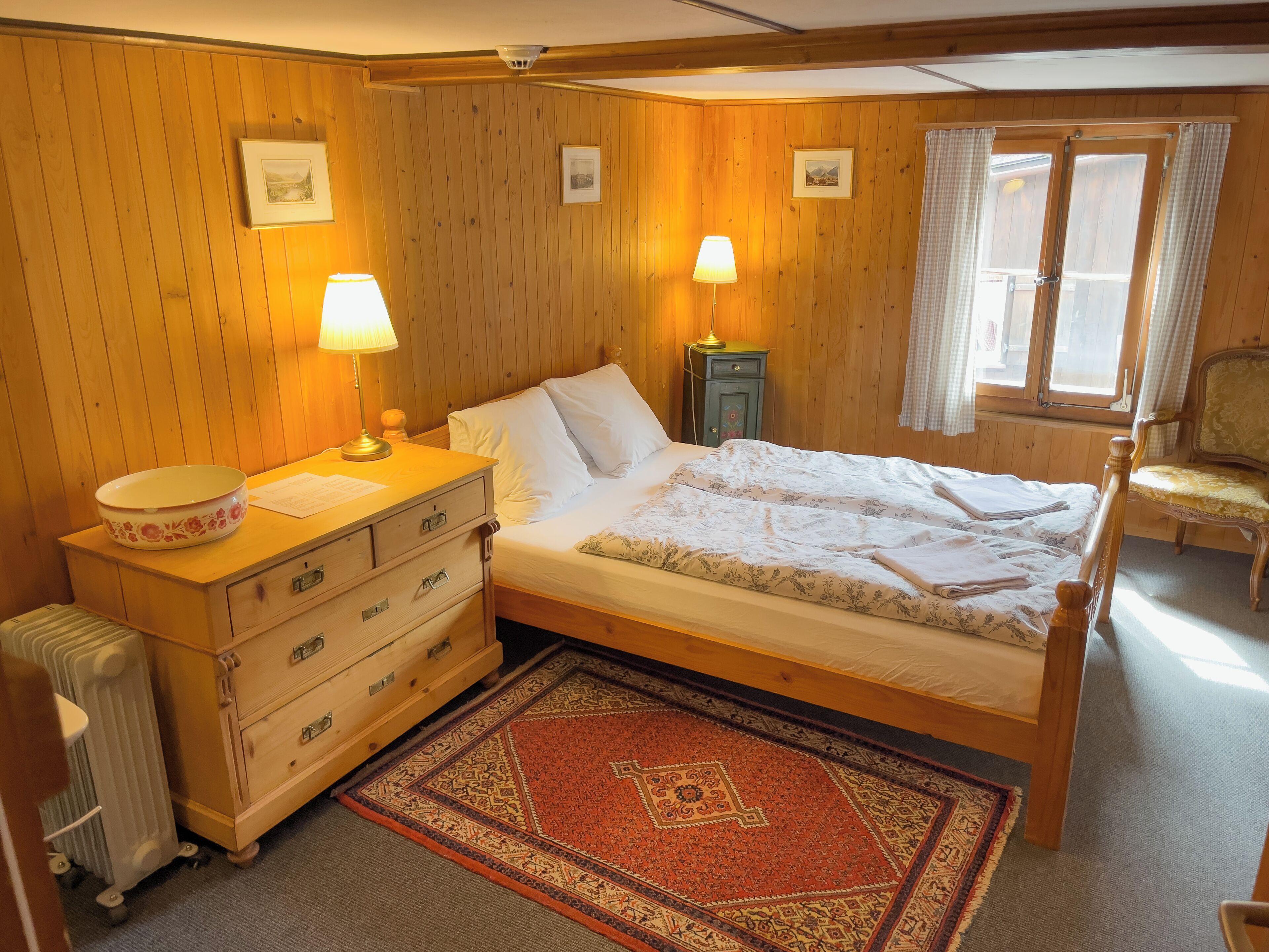 A wooden-paneled bedroom with a bed, two bedside lamps, a dresser, a radiator, a window with curtains, and a chair.