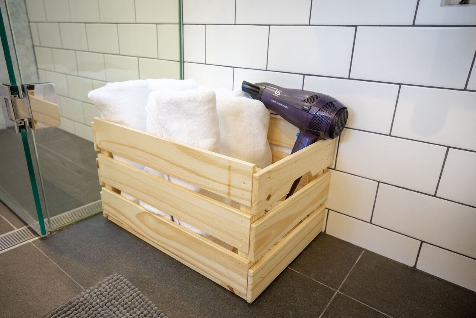 Une caisse en bois contenant des serviettes et un sèche-cheveux, dans une salle de bain dont les murs sont recouverts de carreaux et qui comprend un espace douche.