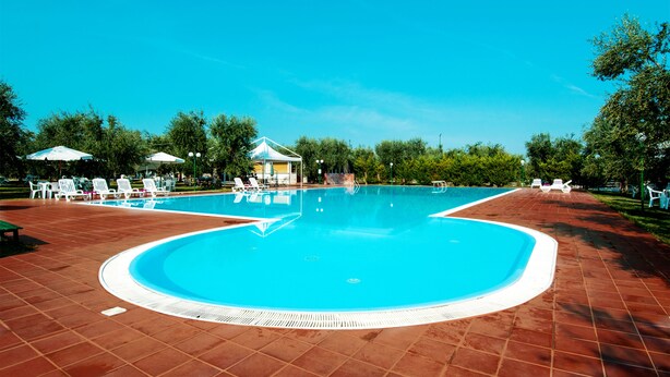 Une piscine entourée d ’arbres, avec des chaises longues blanches et des parasols, ainsi qu’un espace de repos couvert.