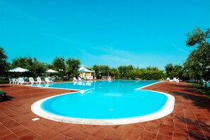 Une piscine entourée d’arbres, avec des chaises longues blanches et des parasols, ainsi qu’un espace de repos couvert.