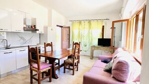 Eine moderne Küche mit weißen Schränken, einem Holzespeisetisch und Stühlen. Im Wohnbereich befindet sich eine rosa Couch und ein Fernseher.