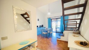 A compact kitchen with a dining area, a staircase, and a large window with blue curtains. - Appartamento Cala D'Ambra (San Teodoro)