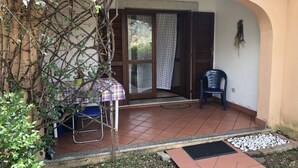 Een overdekt terras met een tafel en stoelen, een deur met een decoratief gordijn, en een kleine tuin.