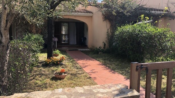 Een pad van stenen leidt naar een huis met een overdekte veranda en een tuin vol potplanten.