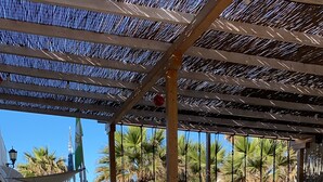 Un café al aire libre con suelo de ladrillo, sillas de metal y una zona con sombra gracias a un techo metálico.