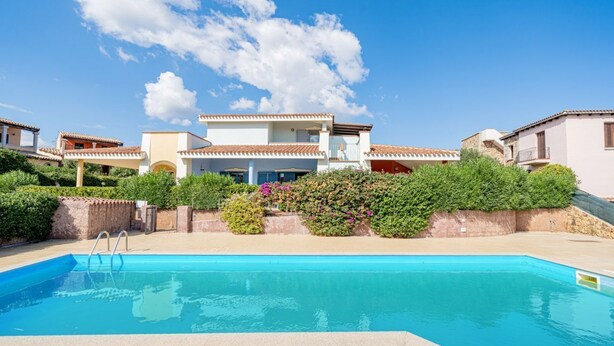 Une piscine à l’eau d’un bleu limpide, entourée d’une terrasse dallée. Au fond, on aperçoit des bâtiments de style architectural méditerranéen, avec des toits en tuiles de terre cuite et des murs en stuc.