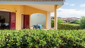 Un bâtiment jaune avec une porte rouge, un patio couvert avec une table et des chaises, et une haie bien entretenue à l’avant.