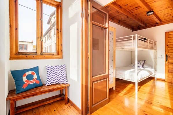 A room with wooden floors, a bench with cushions, a window with a view of buildings, and a bunk bed.