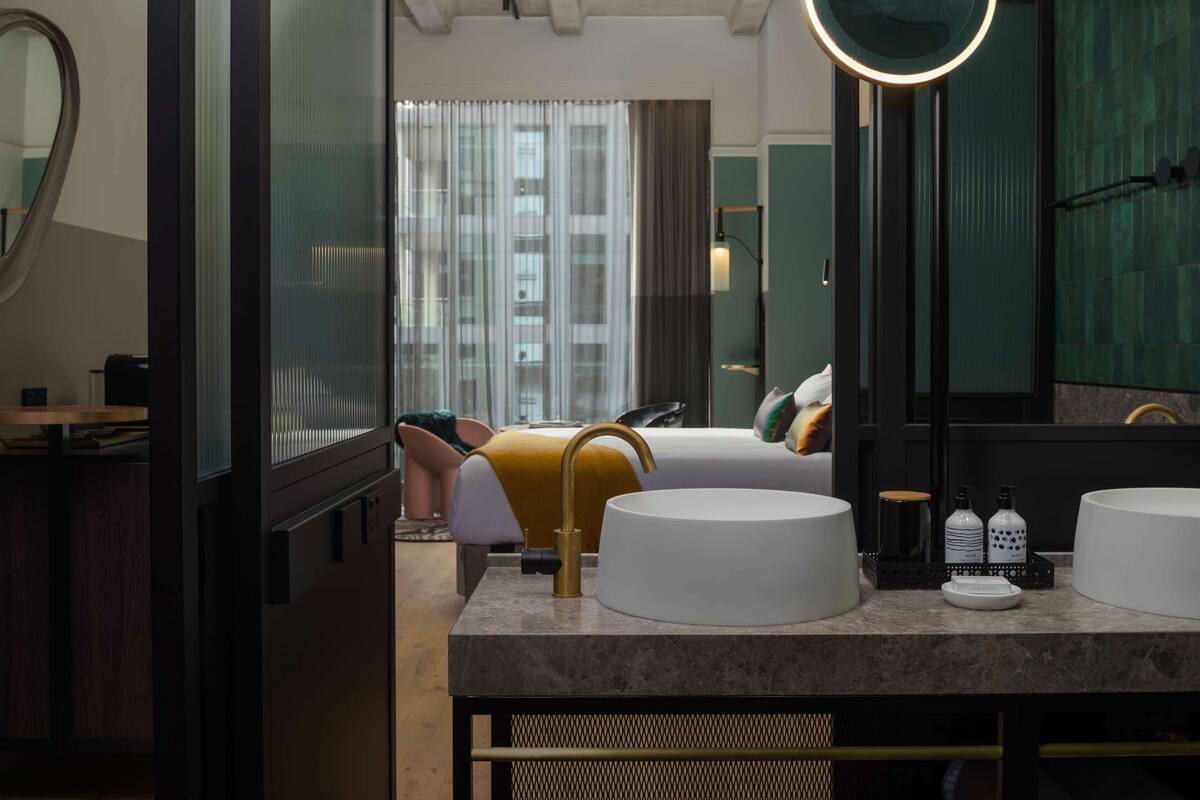 A modern bathroom with a large mirror, two sinks, and a bed in the background.