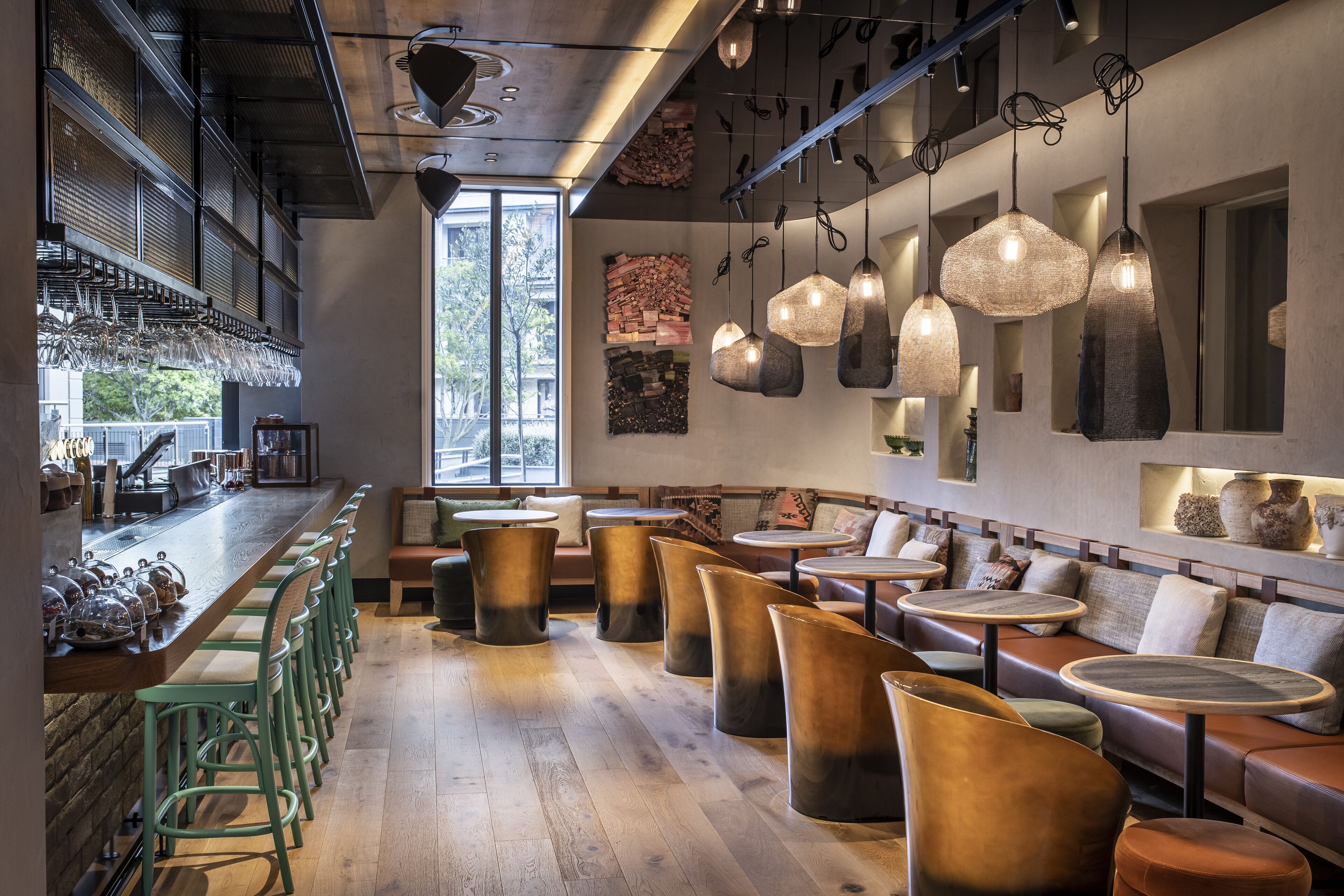 A modern bar area with wooden bar stools, hanging pendant lights, and a large window providing natural light.