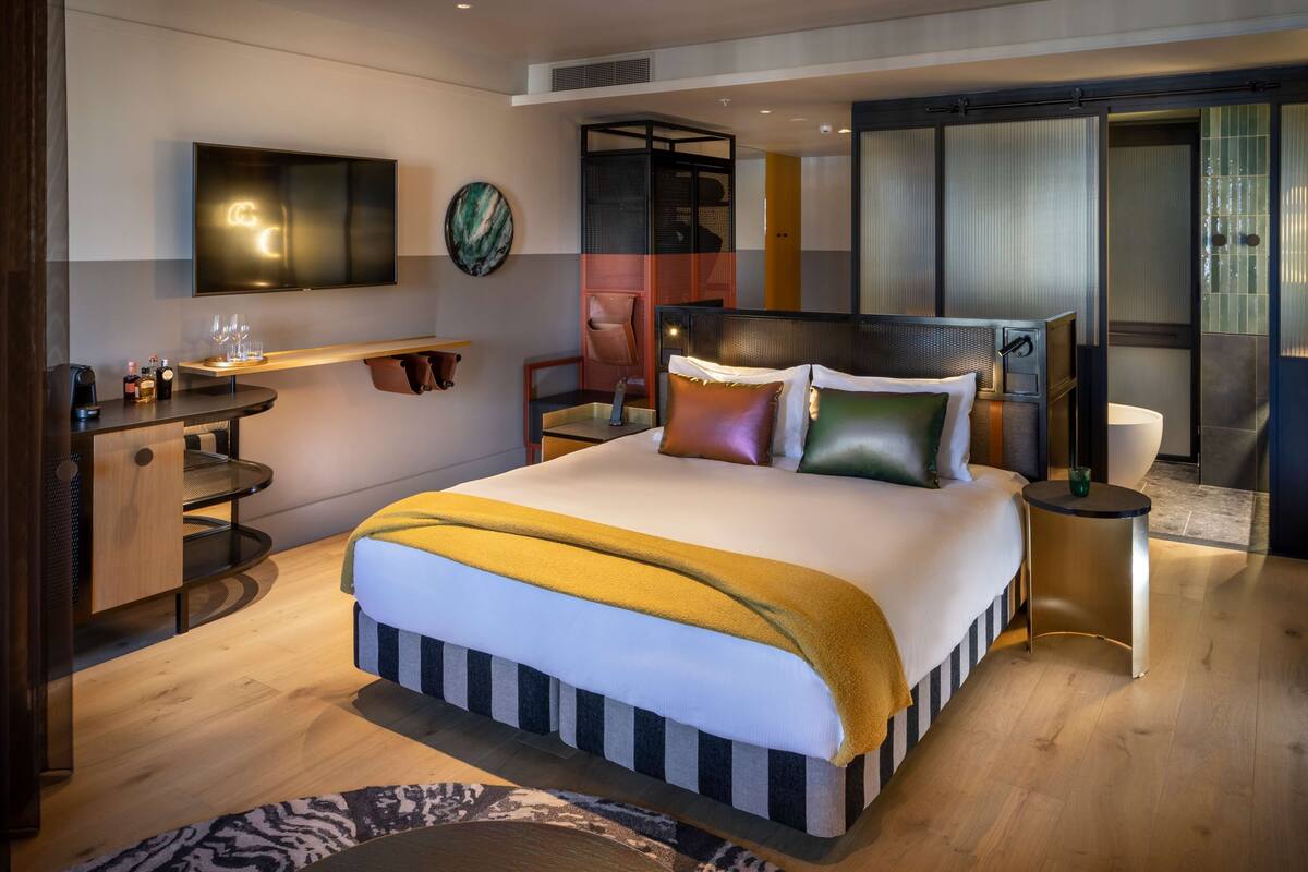 A modern hotel room with a large bed, a desk with a chair, a TV mounted on the wall, and a sliding door leading to a bathroom.