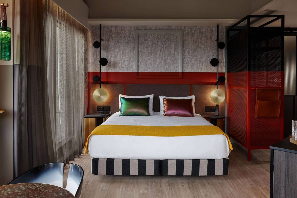 A modern hotel room with a large bed, a red accent wall, and a wooden floor. There is a desk with a chair and a lamp.