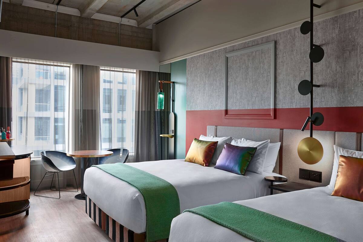 A hotel room with two beds, a desk, a chair, and a view of the city through large windows.
