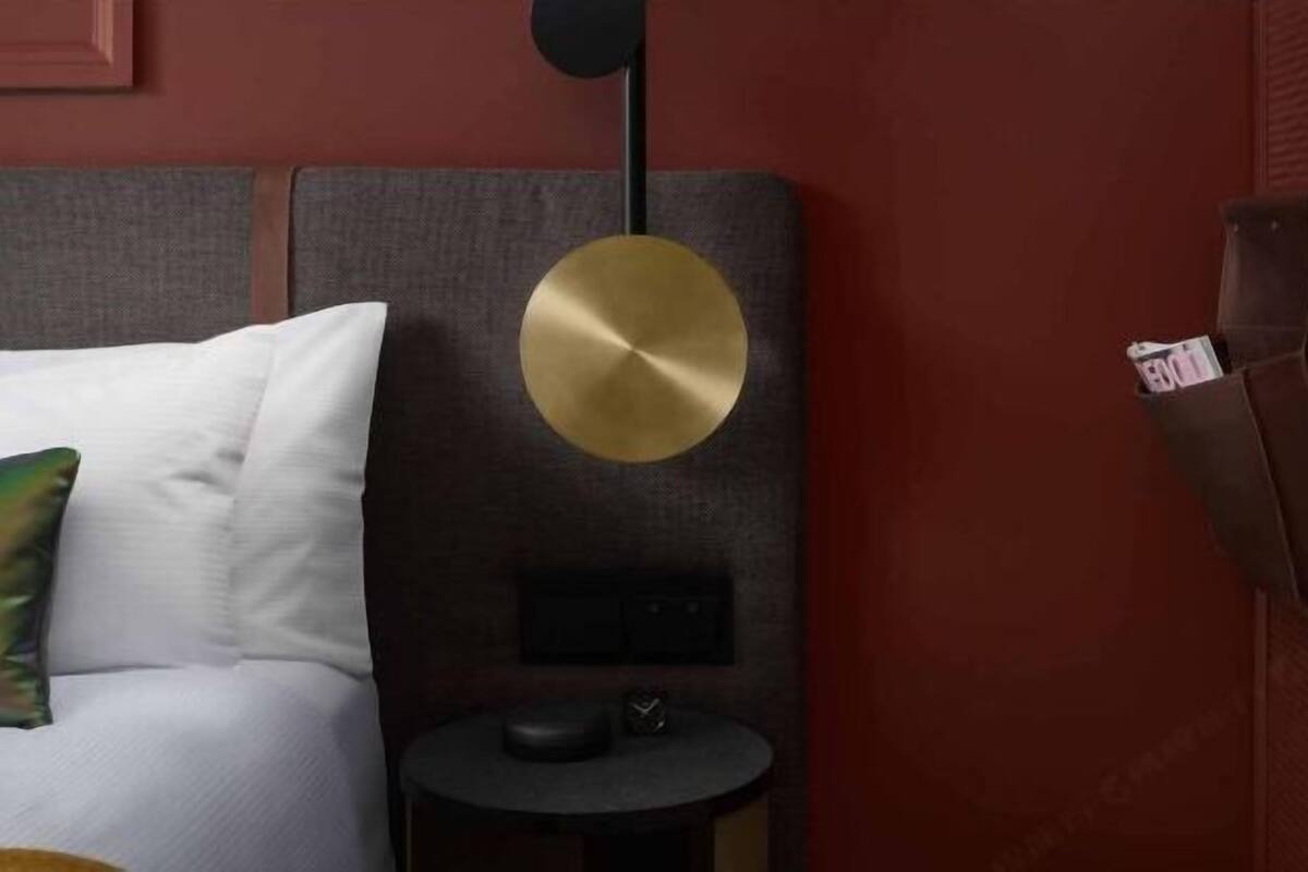 A room with a bed, a small round table, a wall-mounted lamp, and a brown bag hanging on the wall.